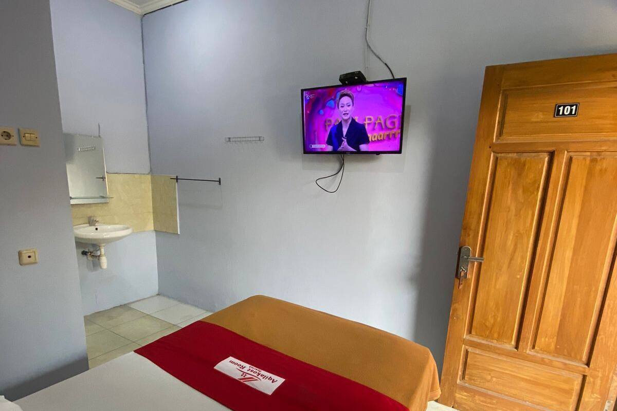 Ein Hotelzimmer mit einem Bett, einem an der Wand befestigten Fernseher, einer Holztür mit einer Nummer sowie einem Badezimmer mit Waschbecken und Spiegel.