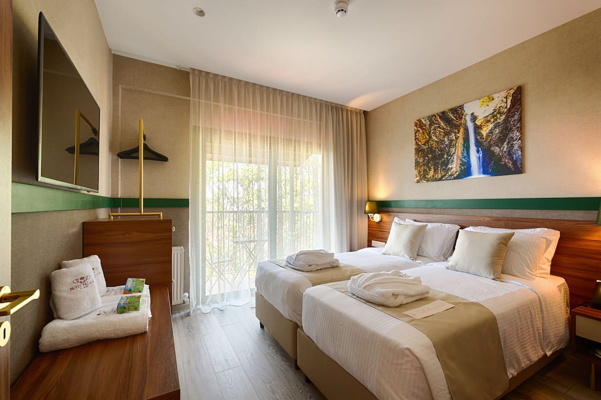 A hotel room with two beds, a large window with sheer curtains, a painting of a waterfall, and a small table with towels and a book.