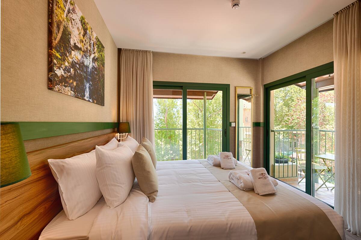 A hotel room with a large bed, a painting on the wall, a balcony with a table and chairs, and a view of greenery outside.