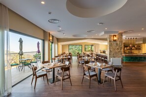 A spacious dining area with multiple tables and chairs, large windows offering views of the outdoors, and a stone accent wall with a built-in bar area. - Petit Palais Platres Boutique Hotel (Platres)