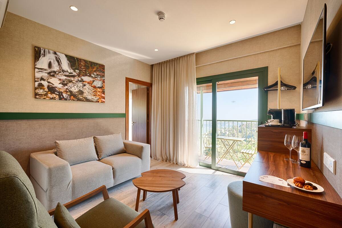A modern hotel room with a sofa, a wooden coffee table, a dining table with a plate of food and wine, and a large window with curtains.