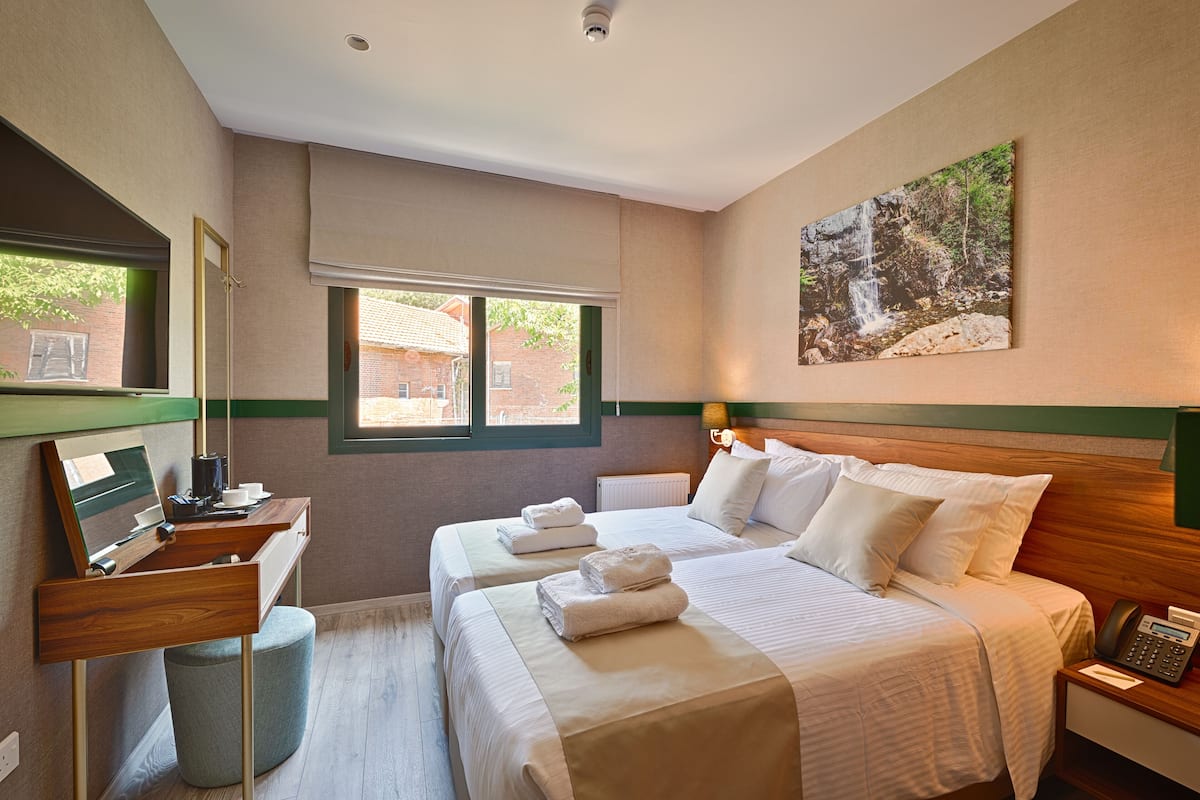 A hotel room with two beds, a desk, a chair, a mirror, a window, and a painting on the wall.