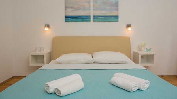 Een net opgemaakt bed met een blauwe spreideken, twee witte kussens en twee opgerolde handdoeken. Twee aan de muur hangende schilderijen beelden een strandscène uit.