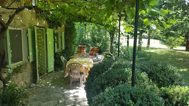 Un área de estar al aire libre techada con mesa y sillas, rodeada de vegetación y un sendero de piedra.
