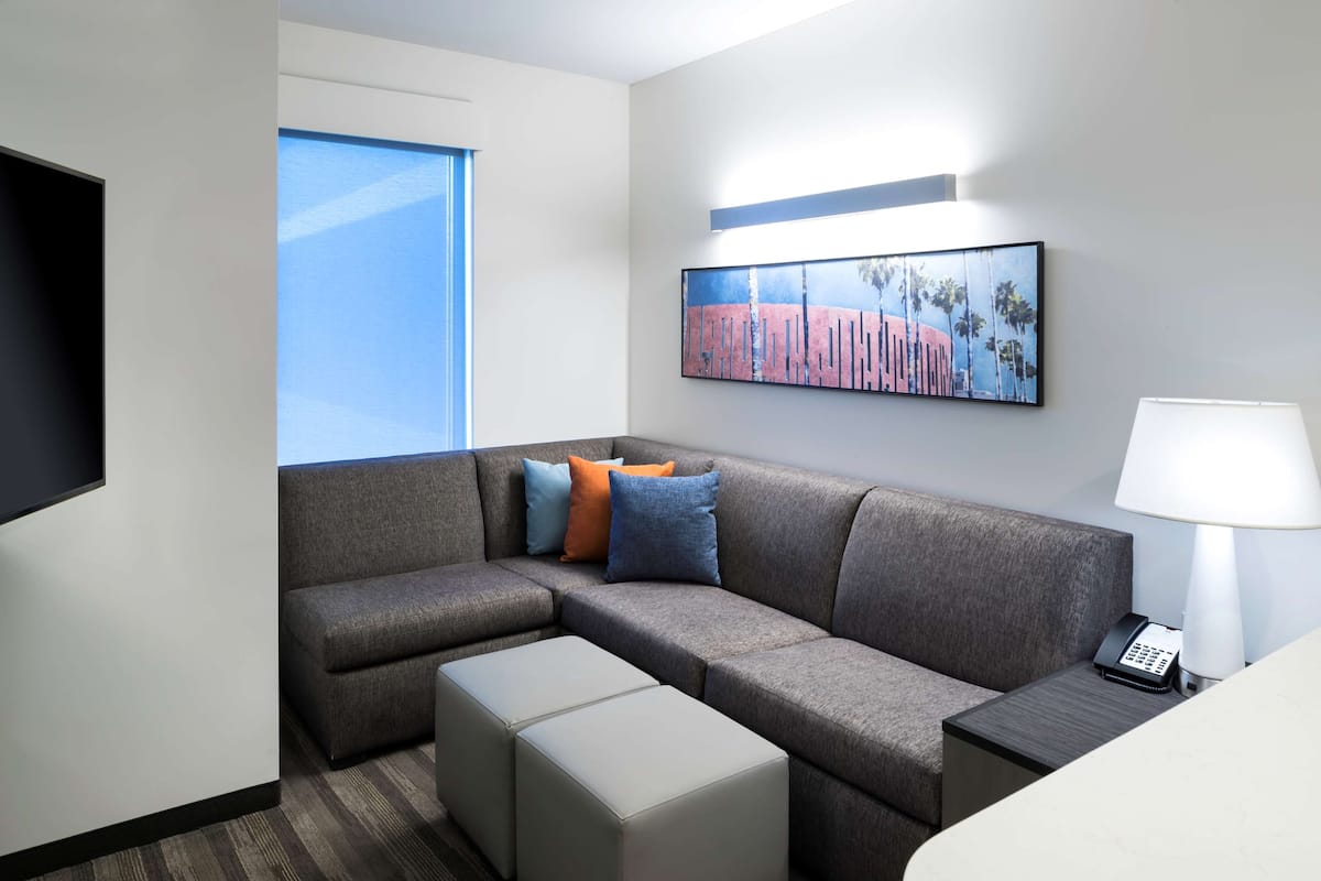 Una habitación de hotel moderna con un sofá modular gris, un televisor de pantalla plana, un cuadro de palmeras, una lámpara y un teléfono sobre una mesita de noche.