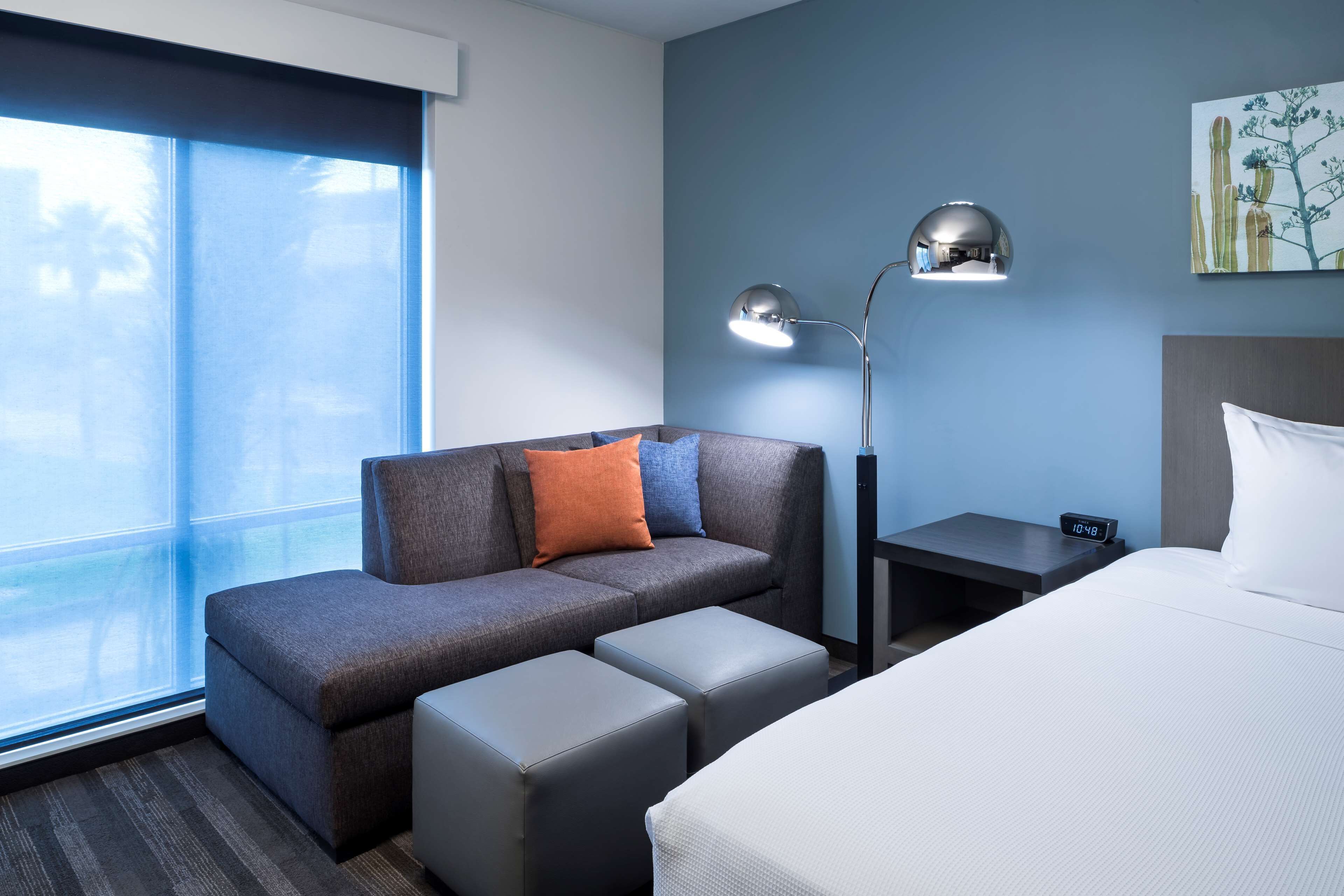 A hotel room with a sofa, ottomans, a bed, a nightstand, a lamp, and a painting on the wall.
