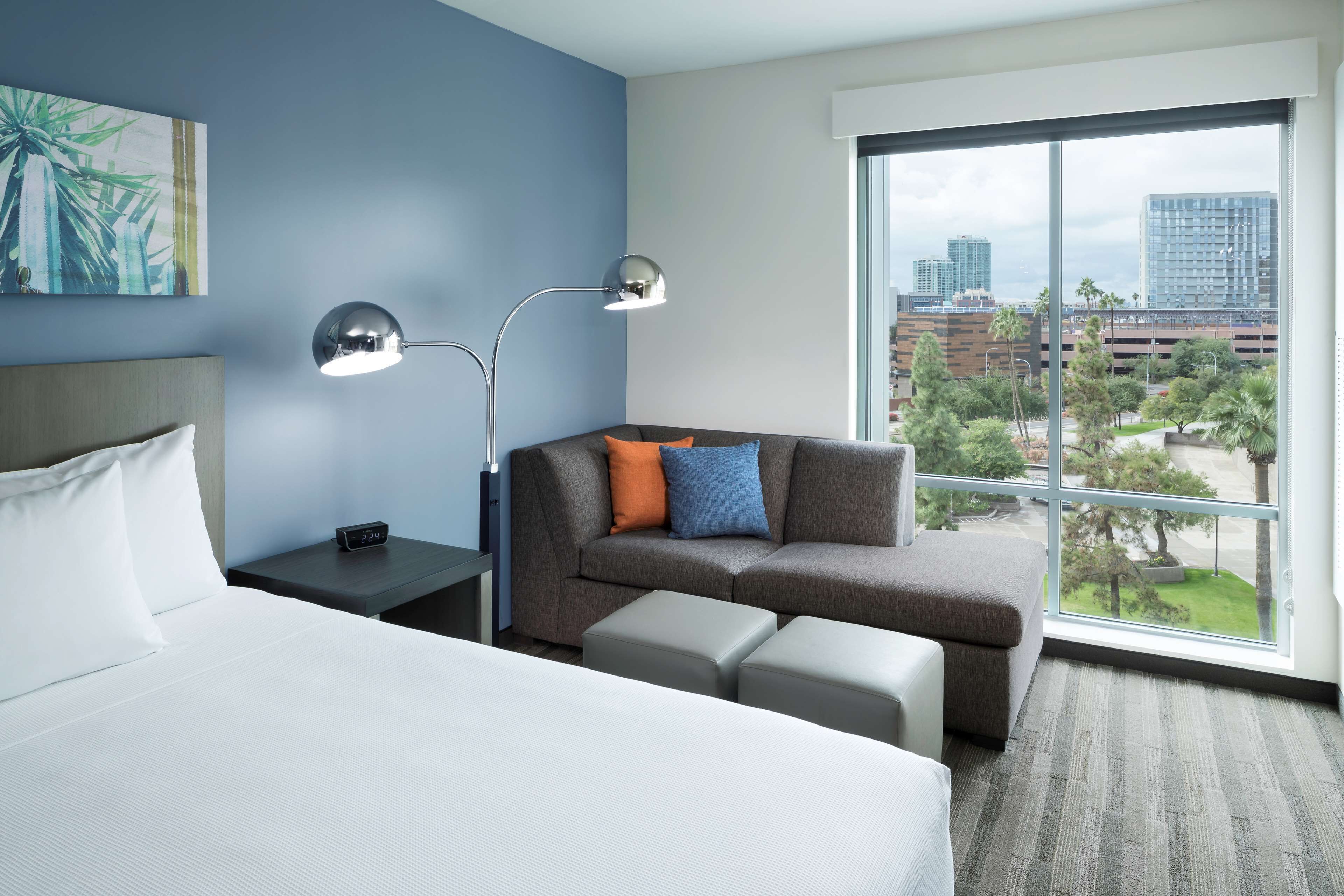 A hotel room with a bed, a gray sofa with orange and blue cushions, a small table with a lamp, and a large window offering a city view.