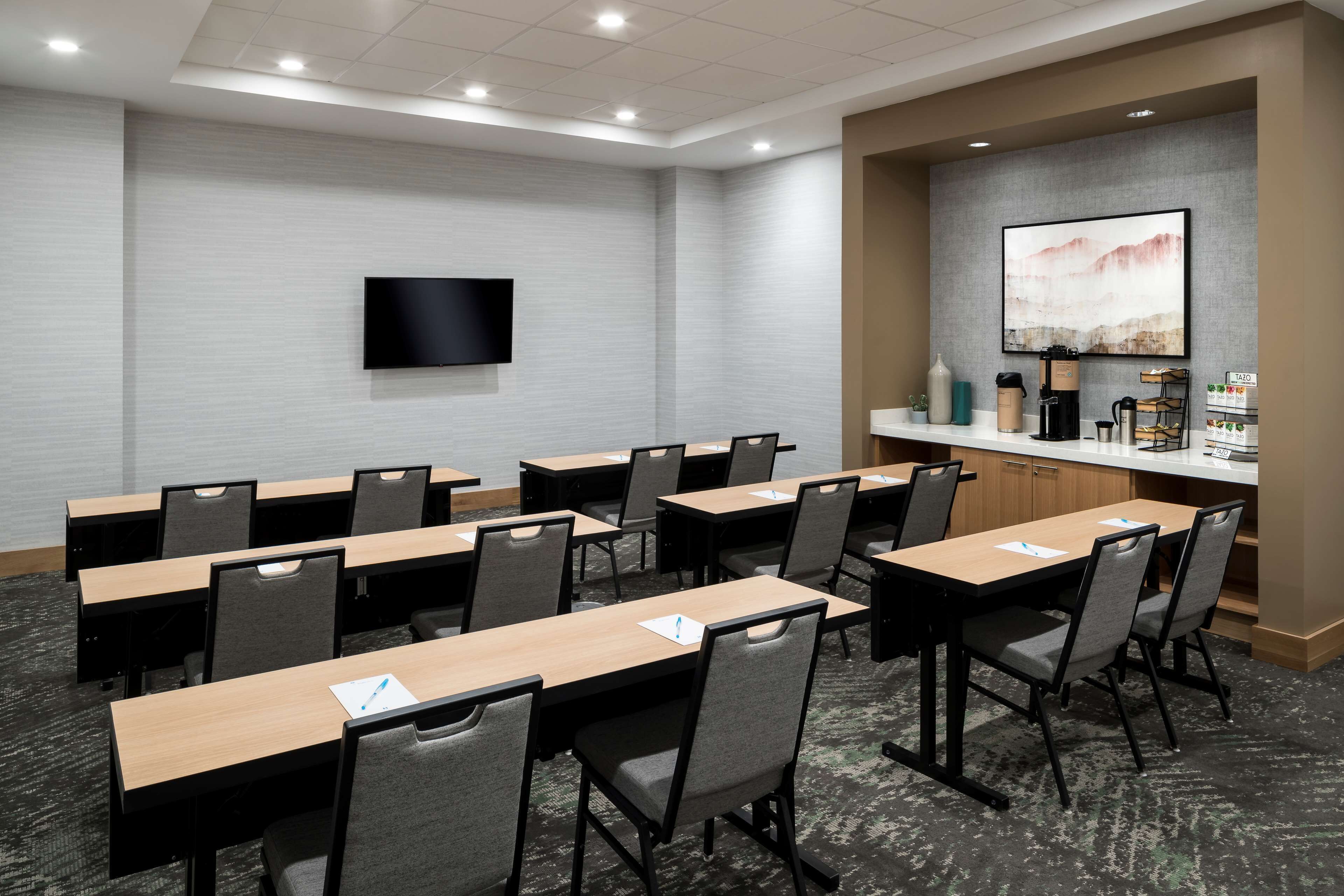 A conference room with multiple tables and chairs, a flat-screen TV mounted on the wall, and a coffee station with various beverages and a coffee maker.