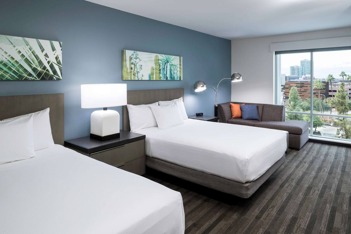 Habitación de hotel con dos camas, un escritorio con lámpara, un sofá y ventanas amplias con vistas a la ciudad.