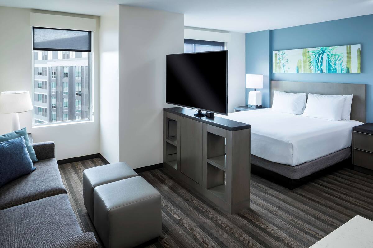 Habitación de hotel con cama, televisión, escritorio, sofá y ventana con vista a la ciudad.
