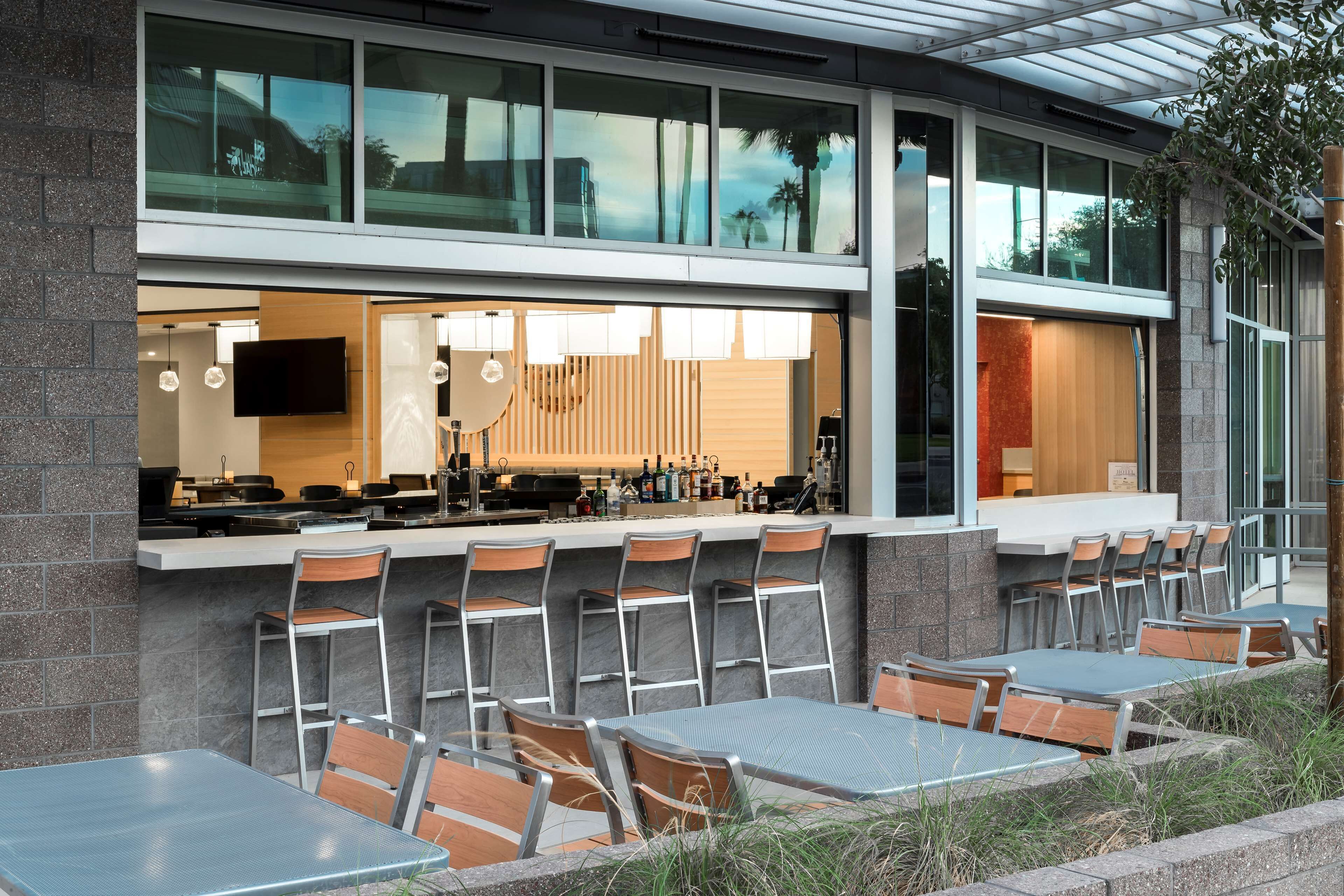 An outdoor dining area with tables and chairs, a bar counter, and a view of the interior through large windows.