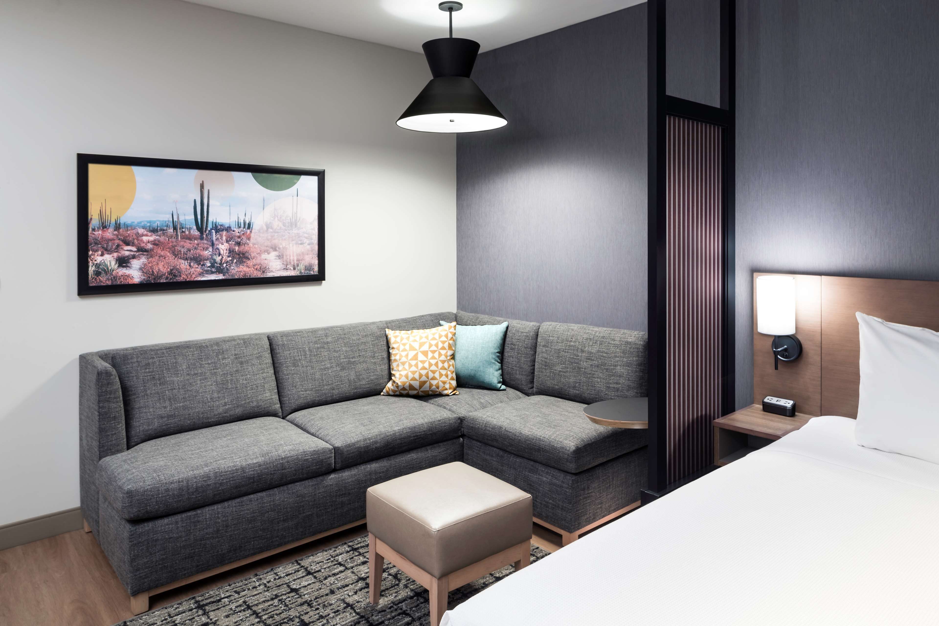Una habitación de hotel moderna con un sofá seccional gris, un pequeño reposapiés, una cama con ropa de cama blanca, una mesita de noche con una lámpara y una obra de arte en la pared que representa un paisaje desértico.