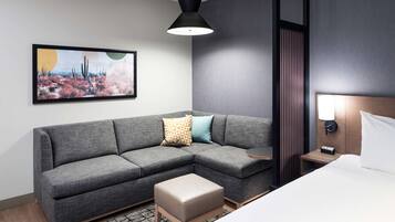 Una habitación de hotel moderna con un sofá seccional gris, un pequeño reposapiés, una cama con ropa de cama blanca, una mesita de noche con una lámpara y una obra de arte en la pared que representa un paisaje desértico.