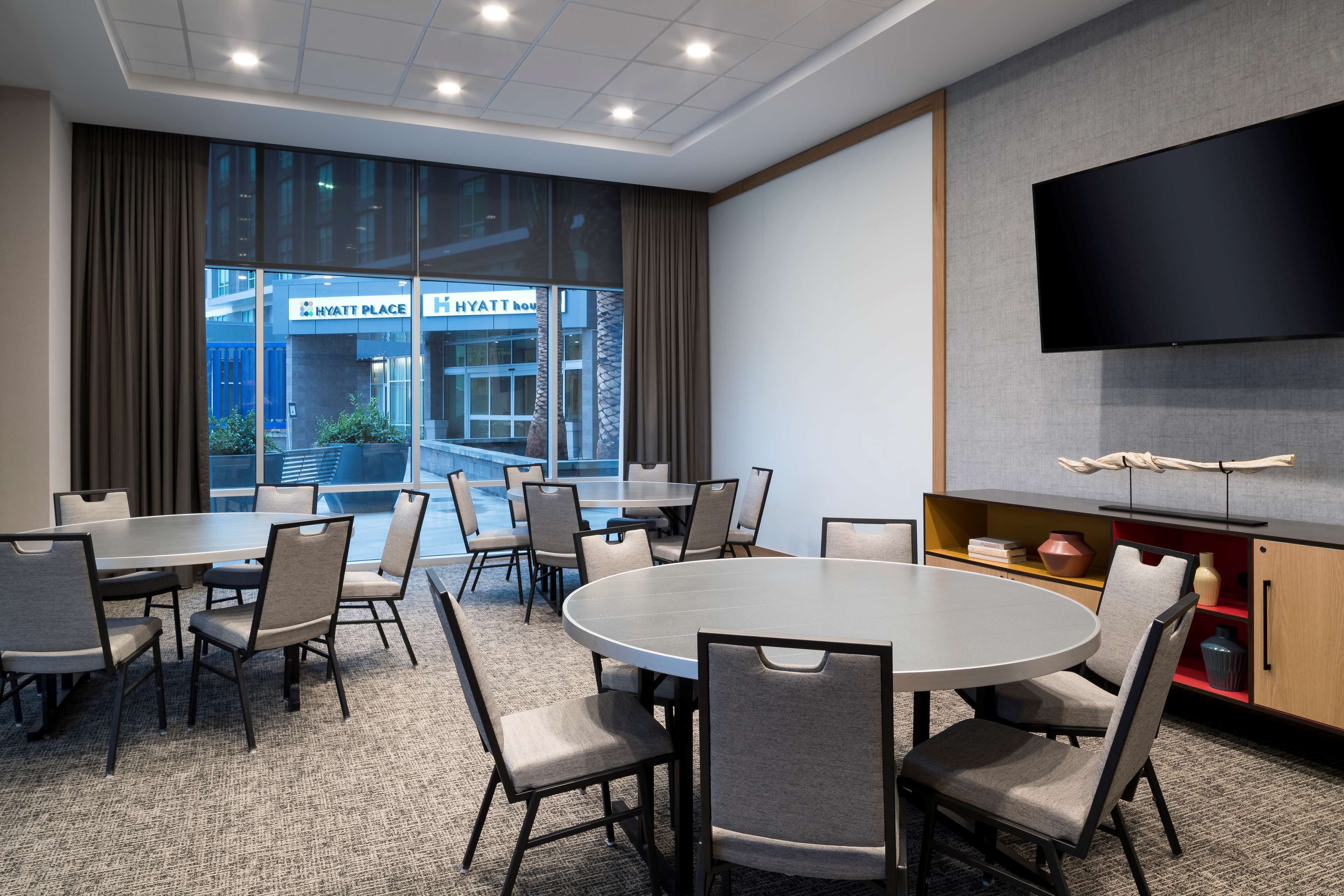 A conference room with a large round table and multiple chairs, a flat-screen TV mounted on the wall, and a view of the Hyatt Place building through large windows.