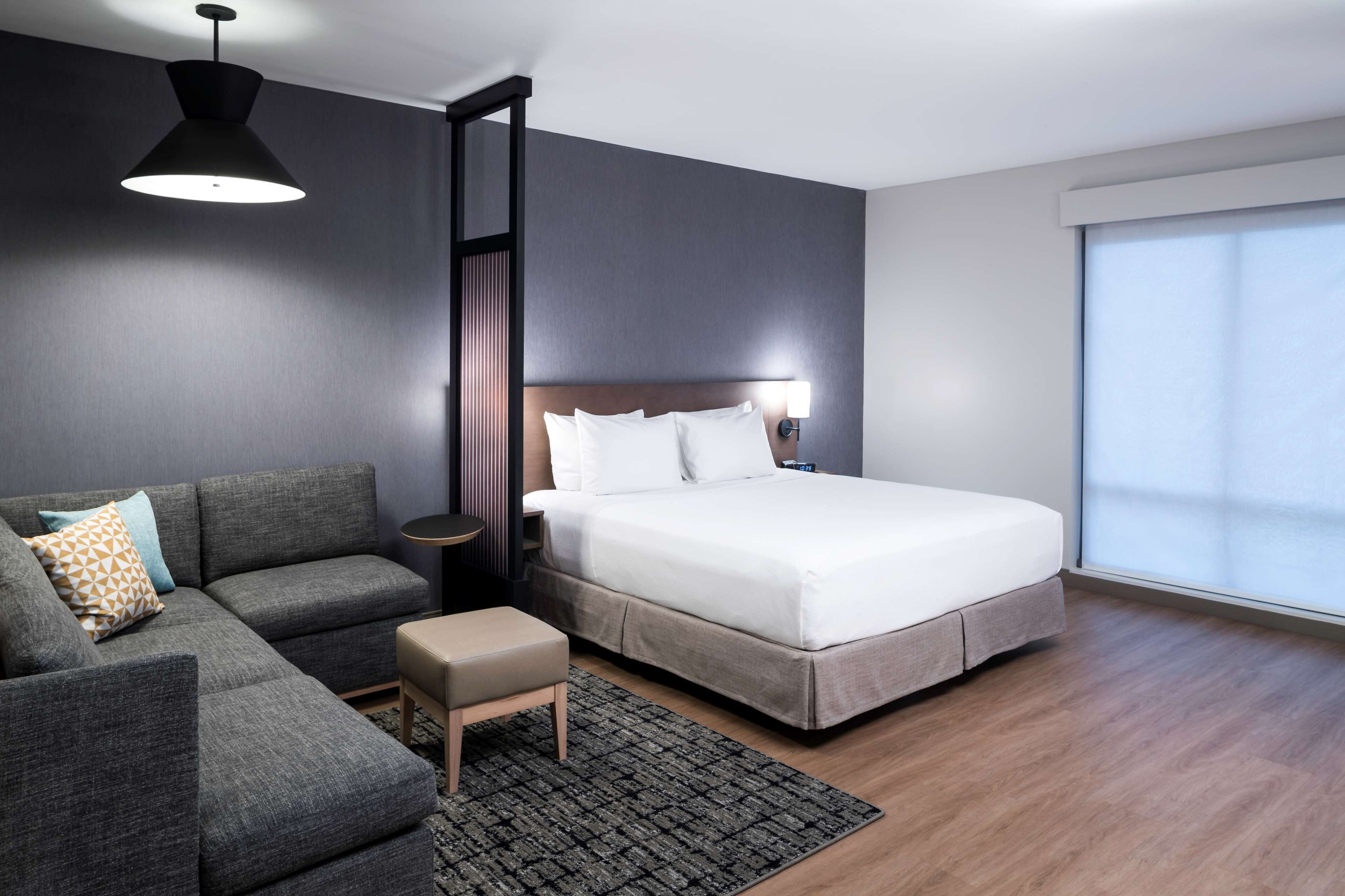 A modern hotel room with a large bed, a sofa, a small table, and a large window.