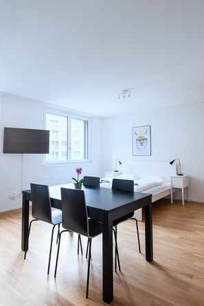 A modern, minimalist room with a large dining table, black chairs, a white bed, a flat-screen TV, and a framed picture on the wall. - Hitrental Messe Apartments (Basel)