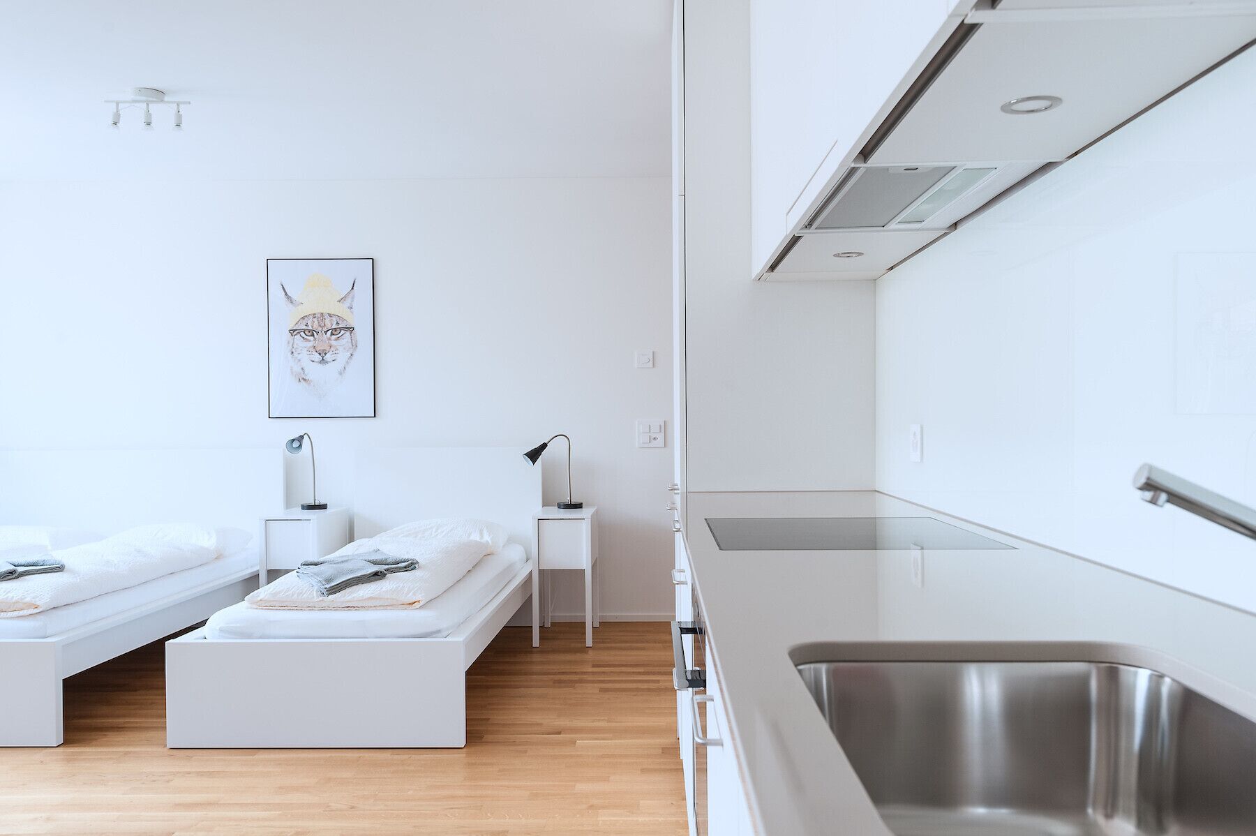 A modern, minimalist bedroom with two single beds, a small bedside table with a lamp, and a framed artwork on the wall.
