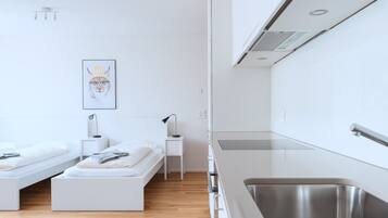 A modern, minimalist bedroom with two single beds, a small bedside table with a lamp, and a framed artwork on the wall.