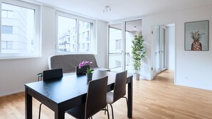 A modern, well-lit room with a dining table, chairs, a potted plant, and a large window offering a view of buildings. - Hitrental Messe Apartments (Basel)