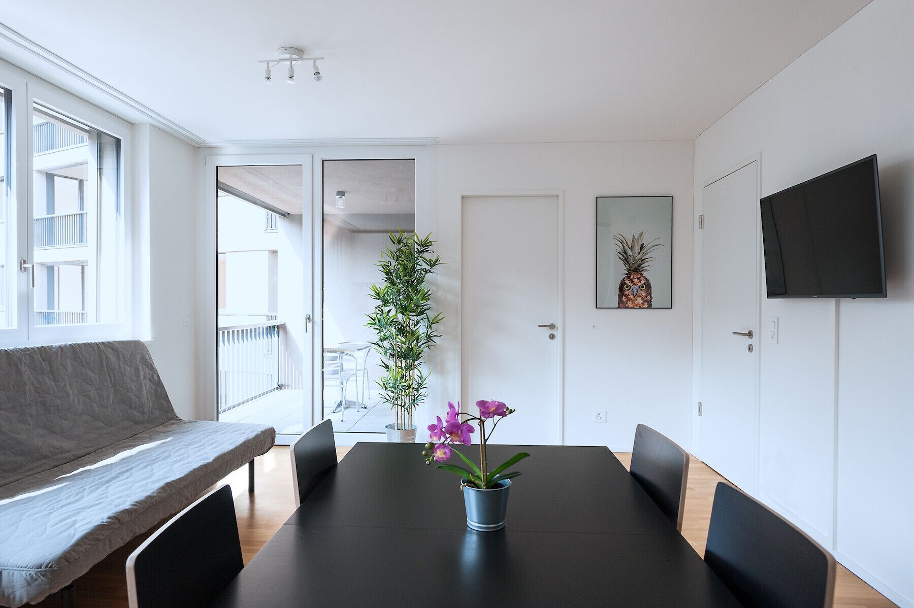 A modern room with a large window, a flat-screen TV, a dining table with chairs, a bench, and a potted plant.