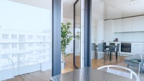A modern kitchen with white cabinets, a dining area with a glass table, and a balcony with a view of buildings. - Hitrental Messe Apartments (Basel)