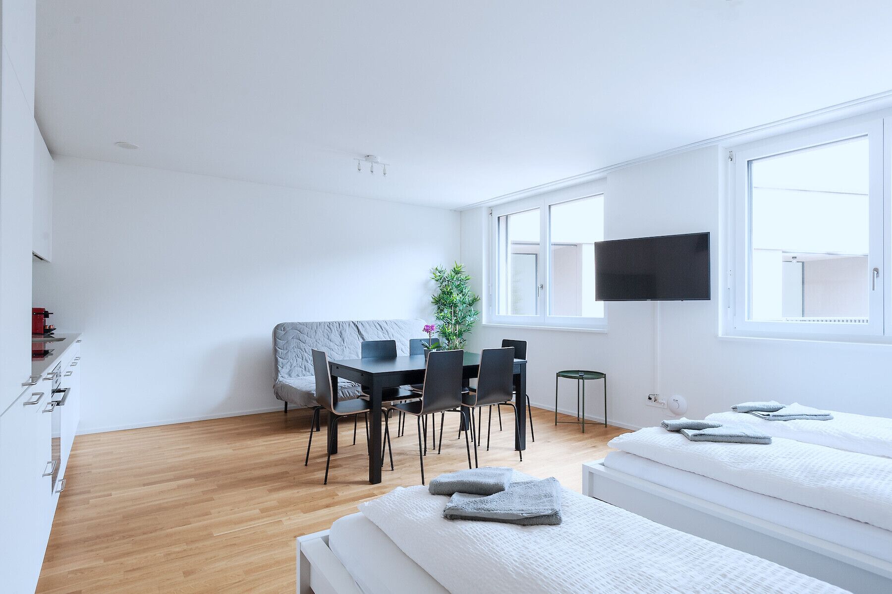 A modern, minimalistic room with a bed, a small dining table with chairs, a TV mounted on the wall, and a window providing natural light.