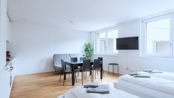 A modern, minimalistic room with a bed, a small dining table with chairs, a TV mounted on the wall, and a window providing natural light.
