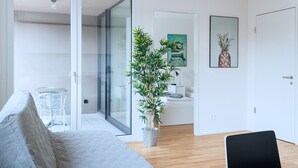 A modern living room with a grey sofa, a black chair, a bamboo plant, and a door leading to another room. - Hitrental Messe Apartments (Basel)