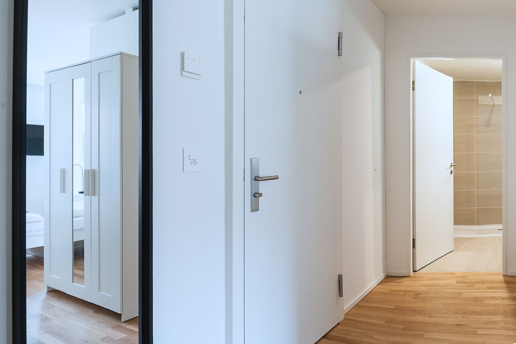 A hallway with a wooden floor, white walls, and a door leading to a bathroom with a shower.
