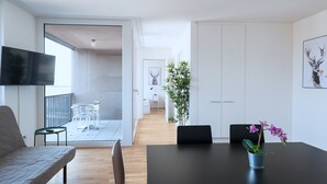 Una sala de estar moderna con un sofá, una mesa de comedor con sillas, un televisor montado en la pared y un ventanal grande.