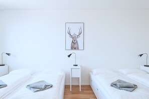 A minimalist bedroom with two beds, a small white table, and a framed deer artwork on the wall. - Hitrental Messe Apartments (Basel)