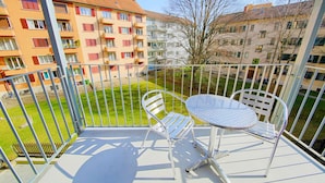 Uma varanda com mesa de metal e duas cadeiras, com vista para um pátio e edifícios ao fundo.