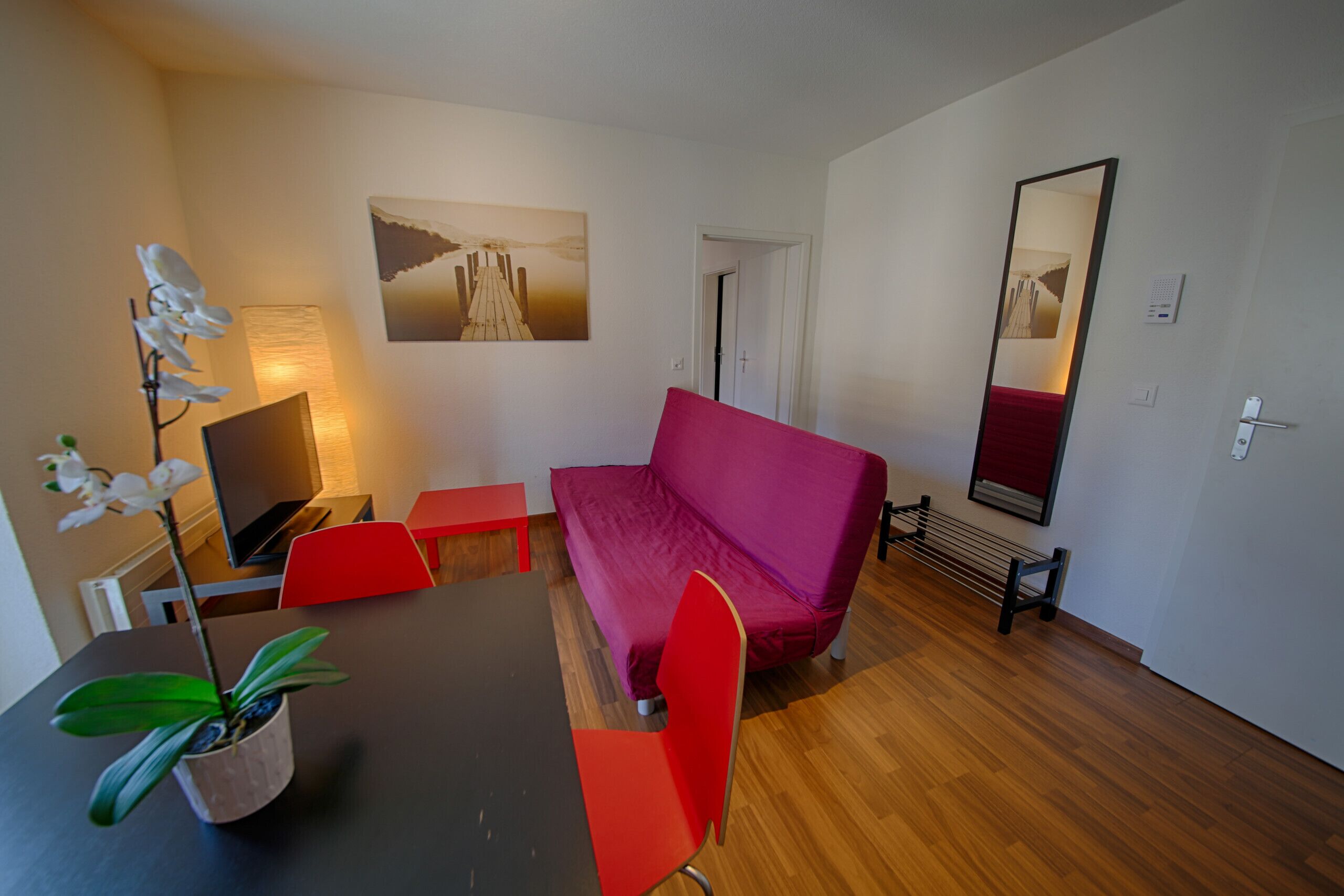 A modern room with a wooden floor, a black dining table, red chairs, a purple sofa, a TV, a lamp, a potted plant, and a painting on the wall.