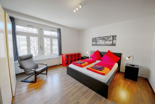 A modern bedroom with a large bed, a chair, a red storage unit, and a window with curtains.