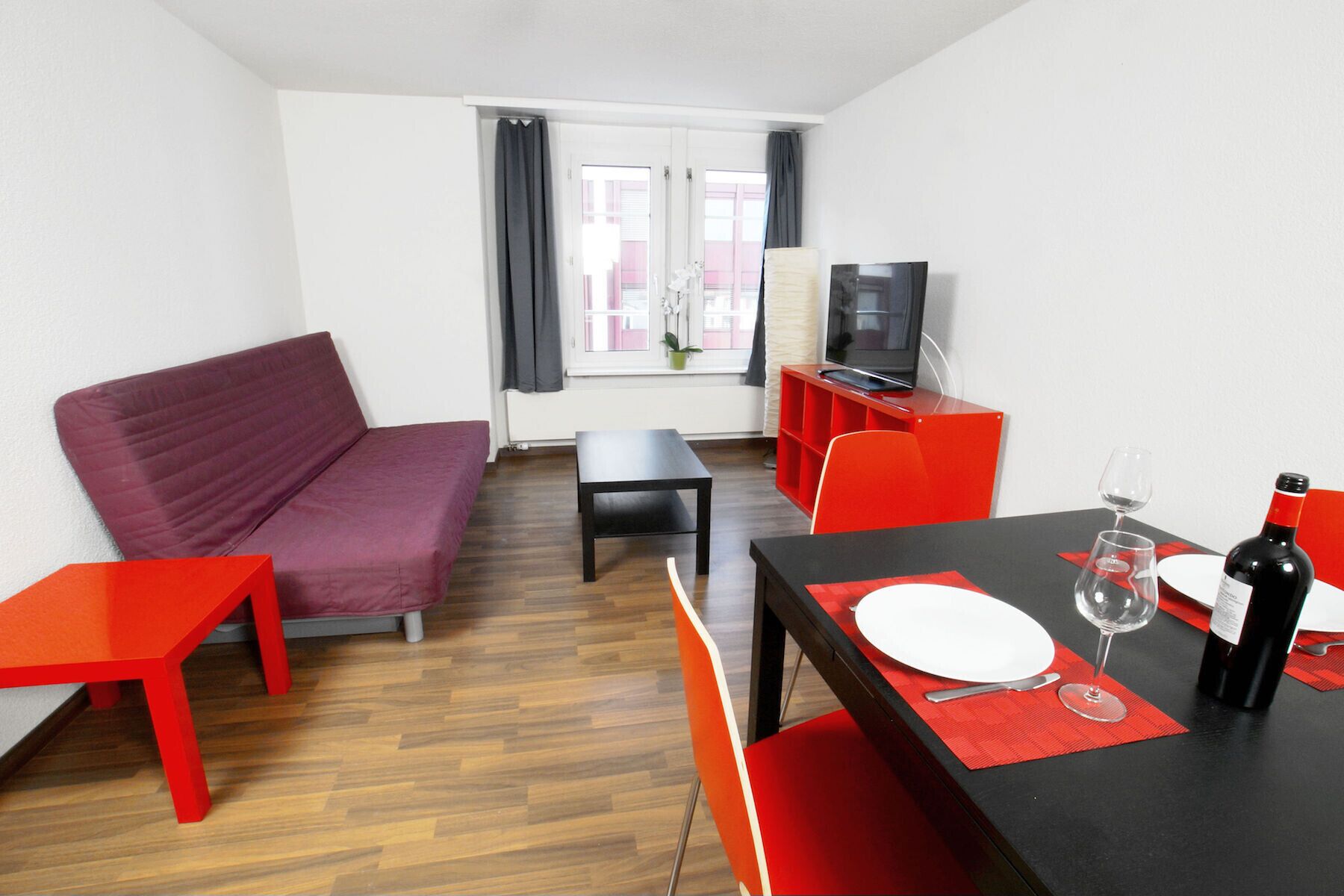 Ein modernes Wohnzimmer mit einem roten Sofa, einem Esstisch mit roten Stühlen, einem Fernseher und einer Weinflasche.
