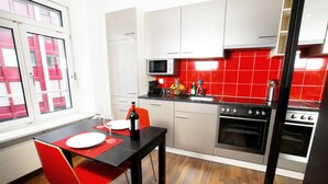 A compact kitchen with a dining area, featuring a red and white color scheme, stainless steel appliances, and a window with blinds. - Hitrental Oerlikon Apartments (Zürich)