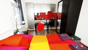 A bedroom with a colorful bedspread, a small dining table with a red chair, and a kitchenette in the background. - Hitrental Oerlikon Apartments (Zürich)