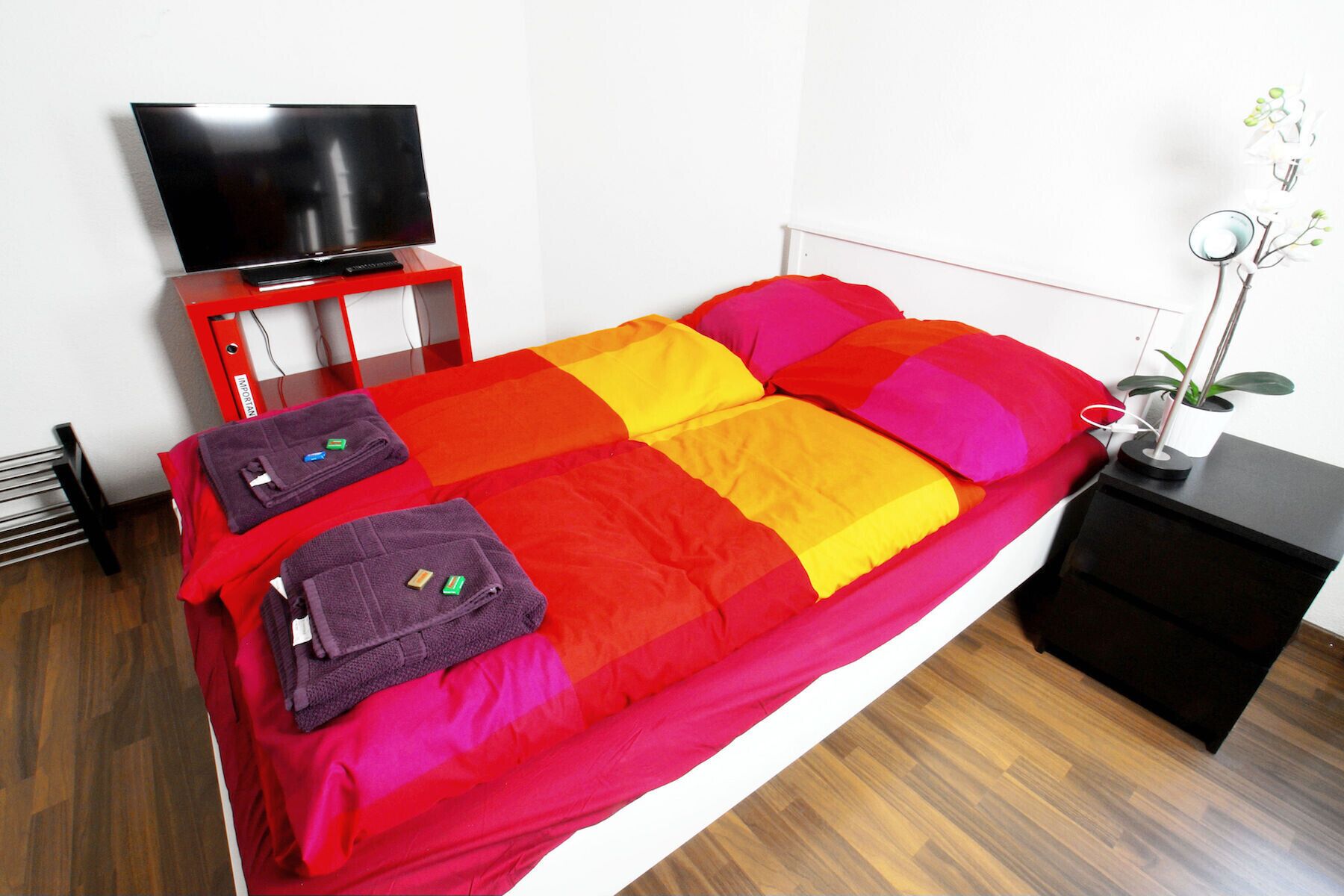 Ein Bett mit bunter Decke, ein Fernseher auf einem Schrank und ein Nachttisch mit Pflanze.