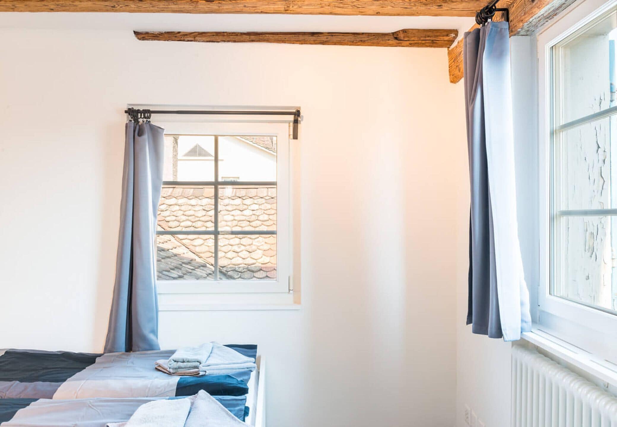 Ein Zimmer mit Bett, einem Vorhangfenster, einem Heizkörper und freiliegenden Holzbalken an der Decke.