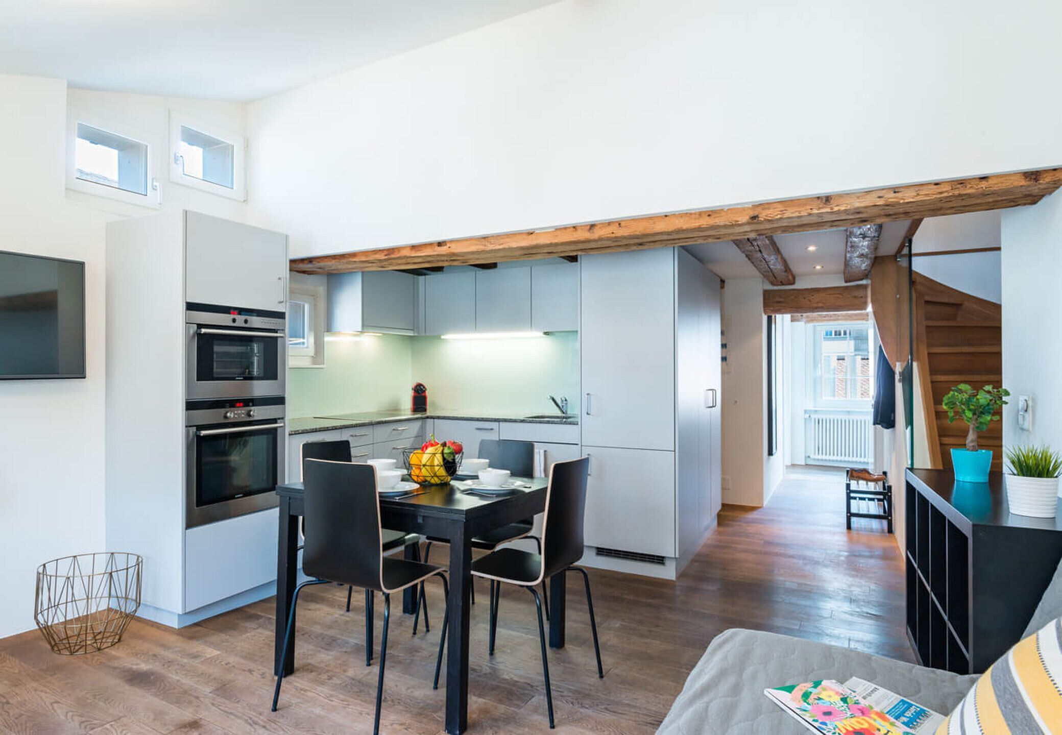 Una cocina moderna con armarios blancos, una zona de comedor con mesa y sillas negras, y una viga de techo de madera.