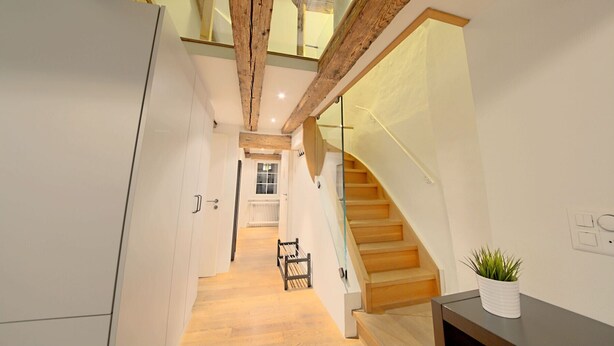 Un couloir étroit avec des poutres en bois, un escalier et une vue sur une pièce où l’on aperçoit un radiateur.