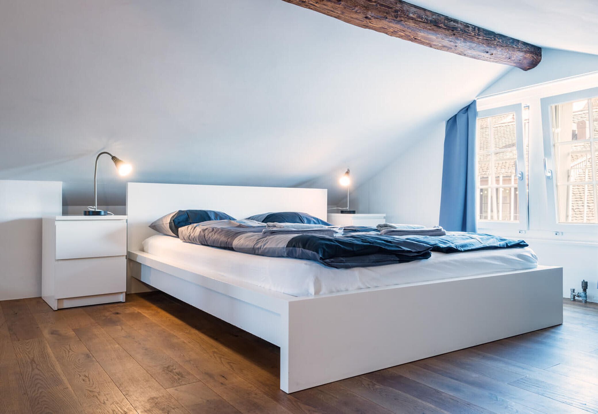 Een moderne slaapkamer met een wit bed, houten vloeren en een schuin dak met zichtbare balken.