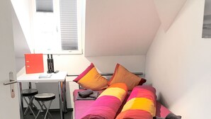 A small, cozy room with a bed, a desk, and a chair. There is a window with blinds, a door, and a small table with a red folder and a bottle. - Hitrental Station Apartments (Lucerne)