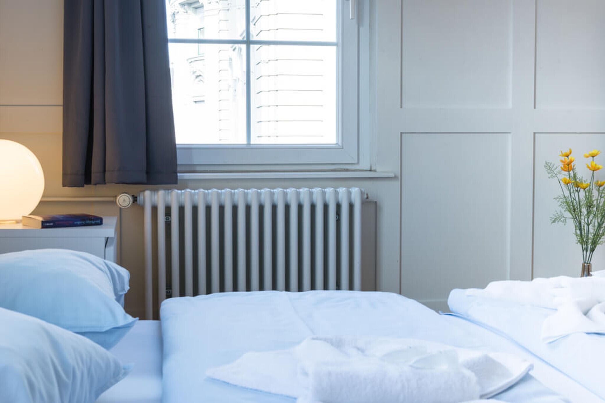A hotel room with a bed, a bedside table with a lamp, a window with curtains, a radiator, and a vase with flowers.
