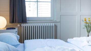 A hotel room with a bed, a bedside table with a lamp, a window with curtains, a radiator, and a vase with flowers.