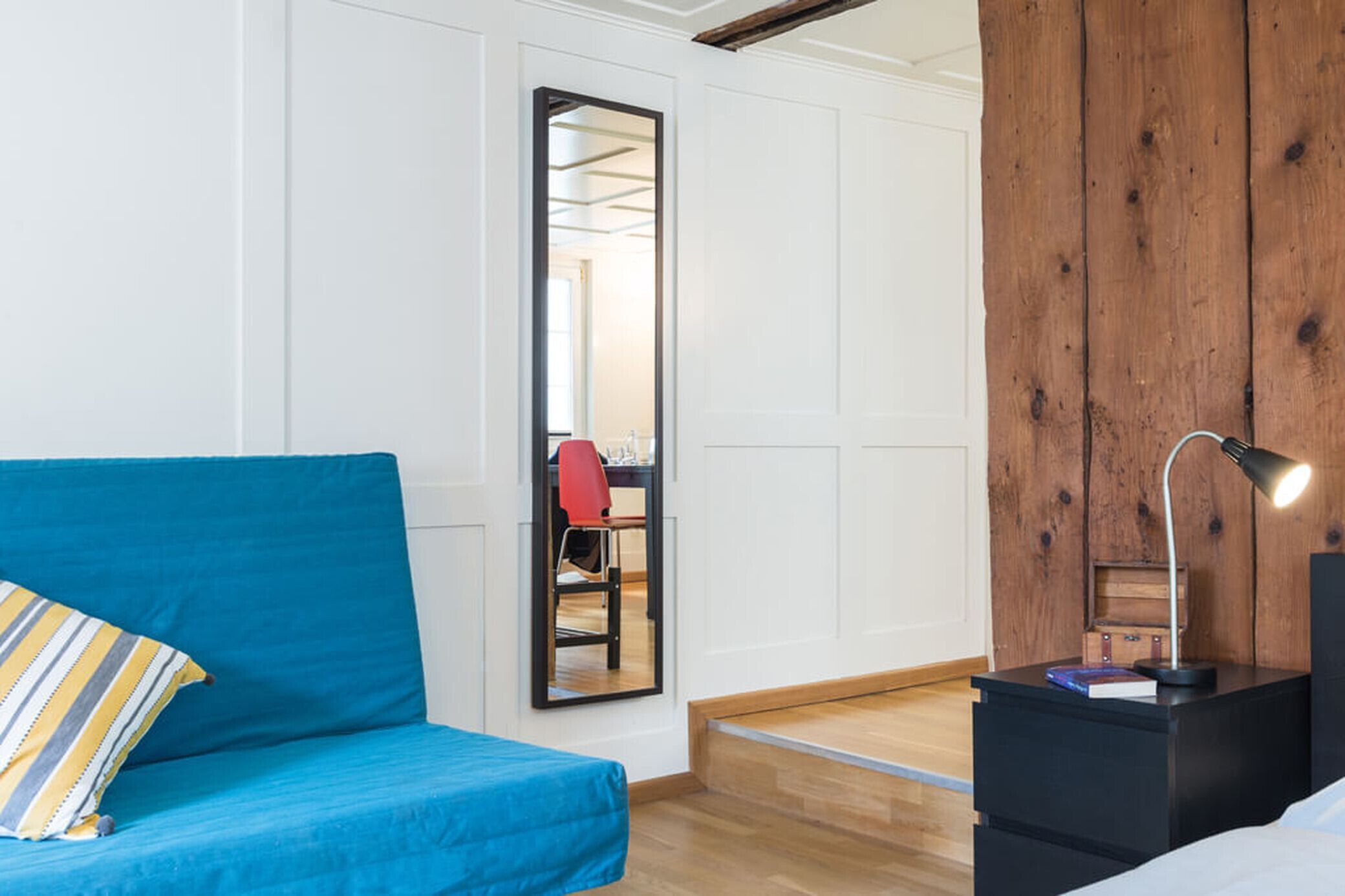 Une pièce avec un canapé bleu, un mur en bois, un miroir, une table de chevet avec une lampe et une petite commode en bois.