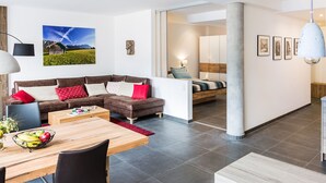 A modern living room with a brown leather sofa, a wooden dining table, and a fruit bowl. There is a large window with a view of mountains and a painting of a mountain landscape. - Exclusive vacation apartment in a unique location in the heart of Mittenwald. (Mittenwald)