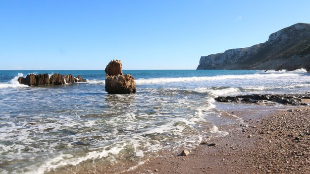 Una costa rocciosa con acque cristalline, una formazione rocciosa imponente e una scogliera sullo sfondo.