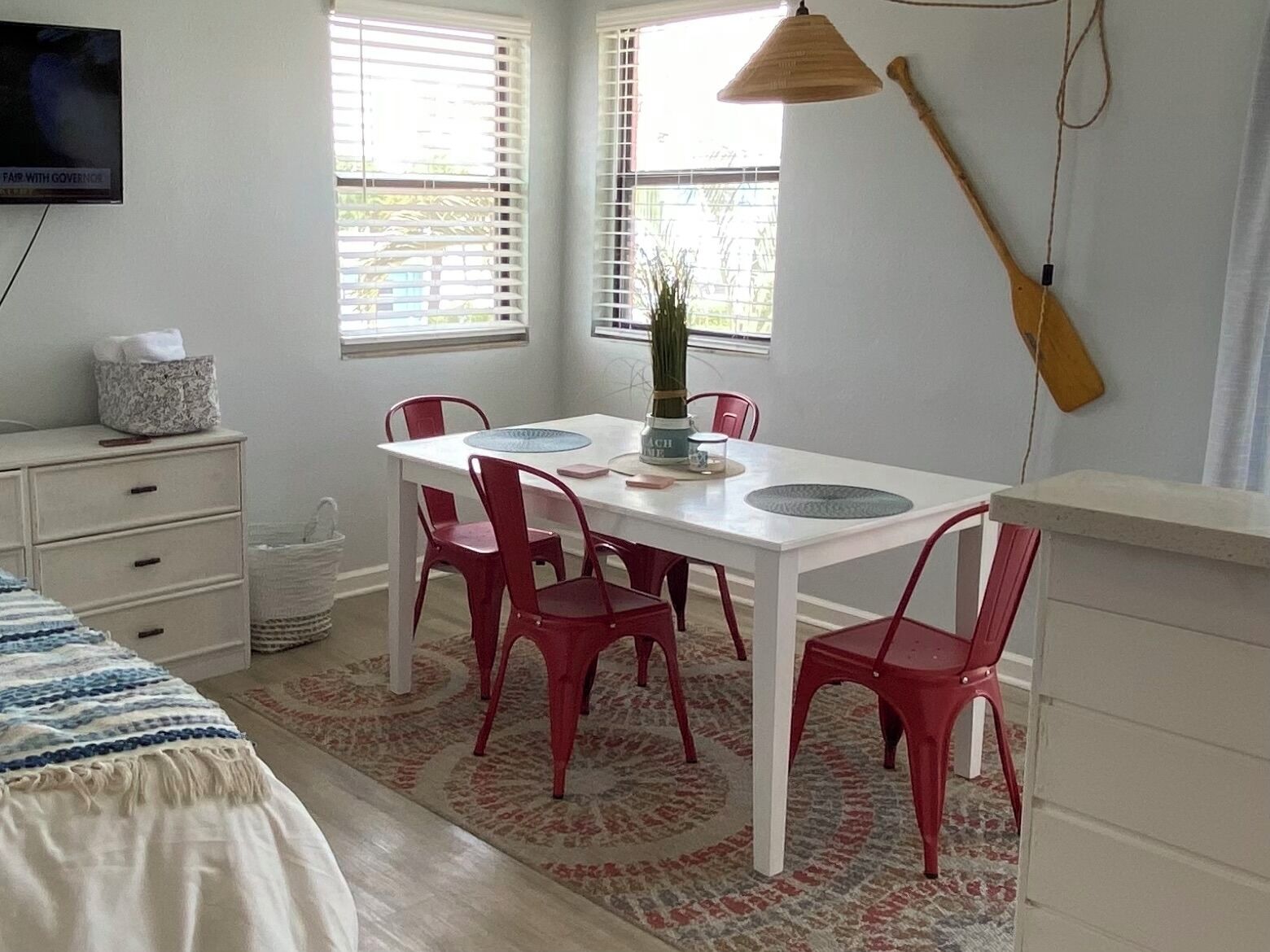 A room with a white table and red chairs, a white dresser, a bed with a blue and white striped blanket, and a wooden guitar leaning against the wall.