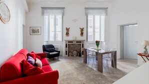A modern living room with a red sofa, a black armchair, and a fireplace. There are two windows with sheer curtains, a dining area with a table and chairs, and a door leading to another room.
